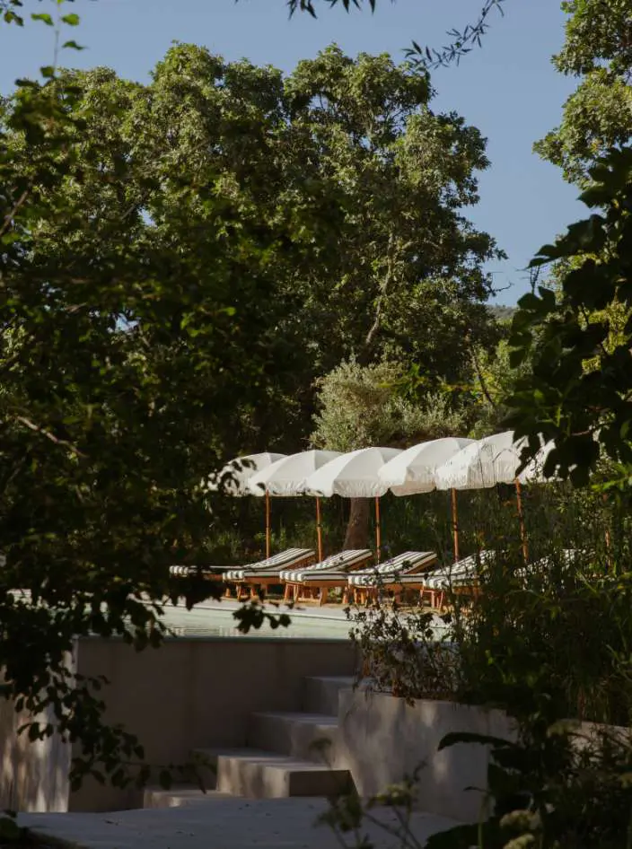 Espreguiçadeiras sob um toldo branco comprido junto à piscina, vistas por entre as árvores