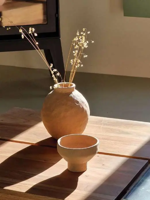 Uma jarra redonda de barro com flores secas e uma taça pequena, numa mesa ao sol