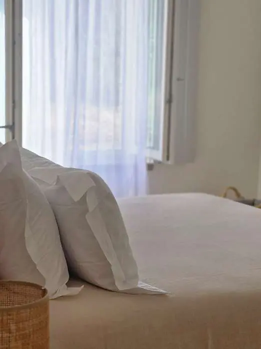 Almofadas brancas numa cama feita, junto a uma janela com cortinado de linho leve