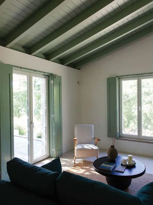 Uma sala sob vigas pintadas de verde, com um sofá verde-escuro virado para as portas abertas